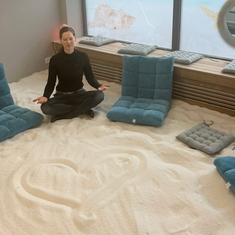 Frau sitzt im Sand, umgeben von blauen Sitzkissen, und meditiert.