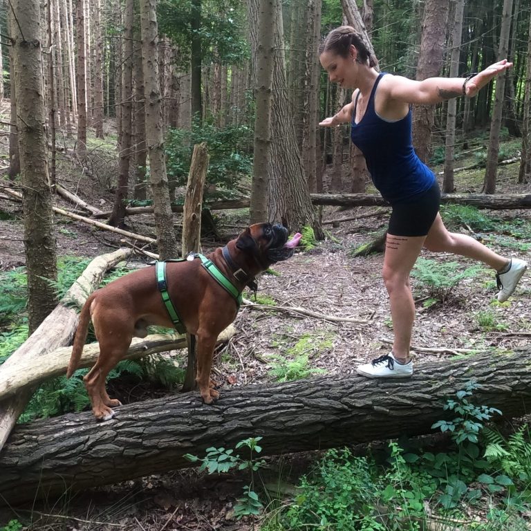 Eine Frau balanciert auf einem Baumstamm, während ein Hund sie aufmerksam anblickt.