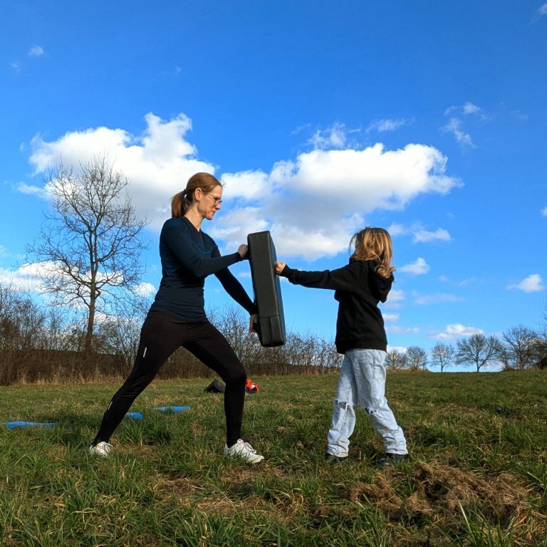 Eine Frau trainiert mit einem Kind auf einer Wiese unter blauem Himmel.
